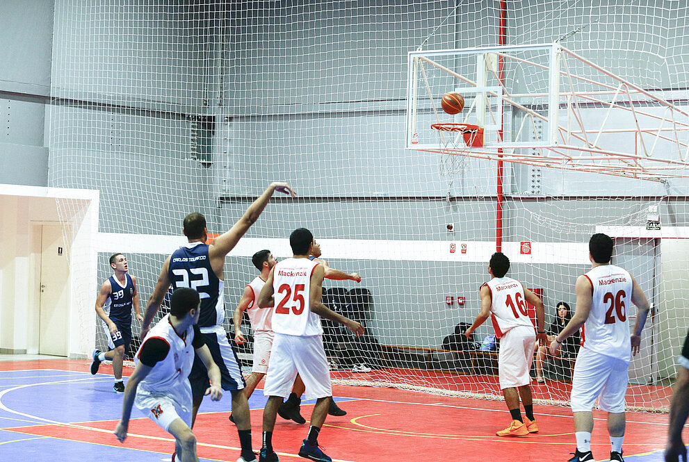 Arremesso pela Escola Naval, com atletas mackenzistas ao redor, olhando a bola indo em direção à rede de basquete.