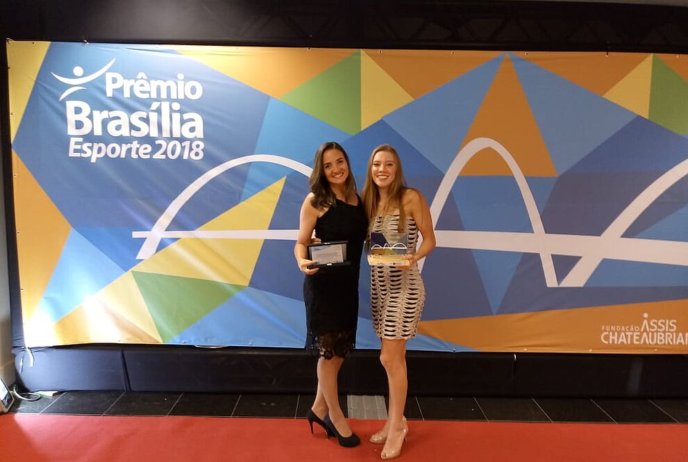 mulheres em vestidos seguram prêmio em frente ao banner do evento