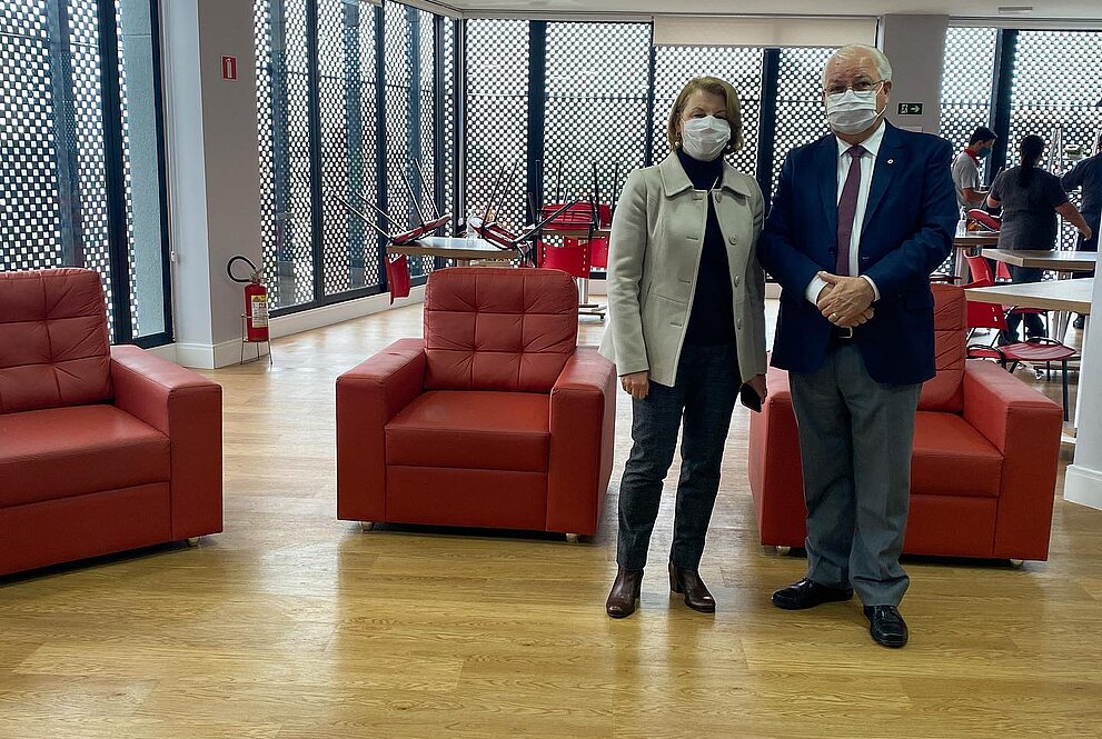 Na imagem, um homem e uma mulher estão de pé em uma sala com poltronas vermelhas e chão de madeira com grandes janelas em torno