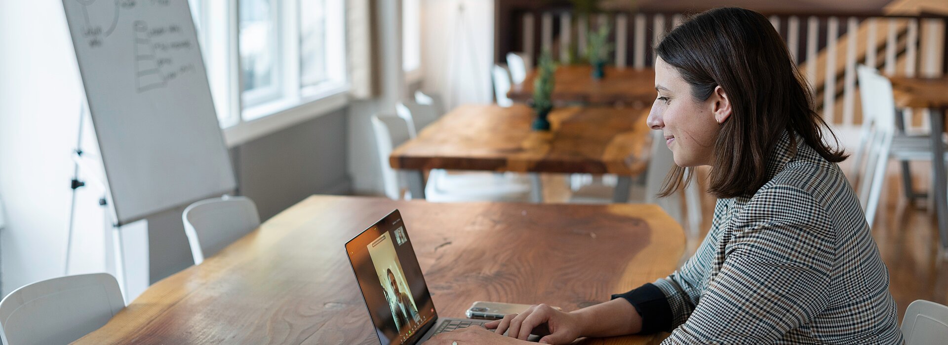 Uma mulher está sentada em uma mesa, em frente a um notebook. O espaço parece ser um café, mas ela está sozinha. Do lado esquerdo da foto podemos ver um quadro, com algumas coisas escritas. 