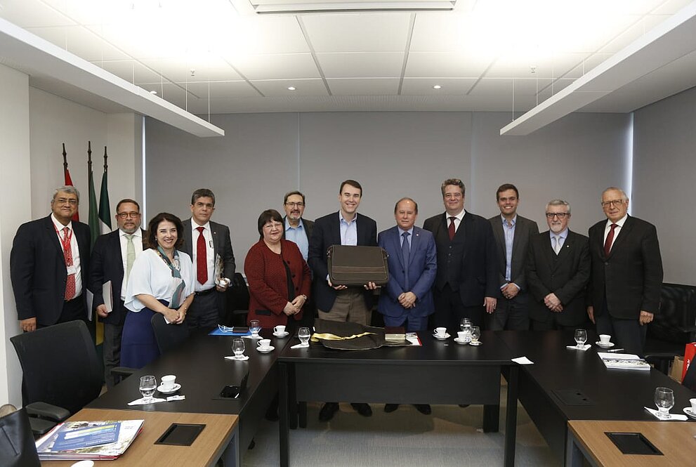 Autoridades mackenzistas ao lado dos representantes da Gordon College, 12 pessoas de pé em frente à mesa de reunião em uma sala com fundo branco.
