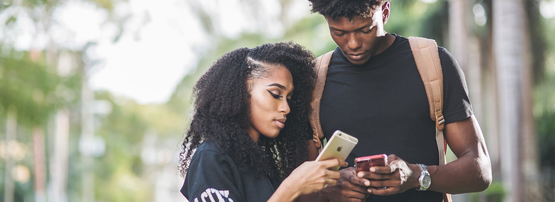 Na imagem, duas pessoas conferem um celular. Ao fundo, pode-se ver árvores e parte do céu