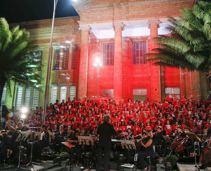 Imagem mostra coral do Mackenzie durante cantata de Natal realizada em 2018. A foto tem árvores, estrutura do seminário de tijolinhos, luz vermelha e o coral. 