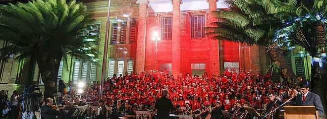 Imagem mostra coral do Mackenzie durante cantata de Natal realizada em 2018. A foto tem árvores, estrutura do seminário de tijolinhos, luz vermelha e o coral. 