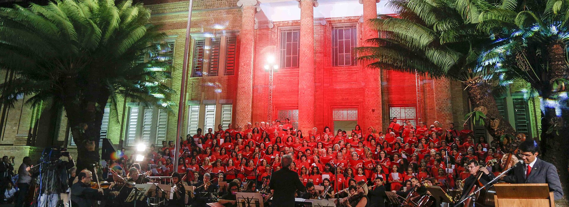 Imagem mostra coral do Mackenzie durante cantata de Natal realizada em 2018. A foto tem árvores, estrutura do seminário de tijolinhos, luz vermelha e o coral.
