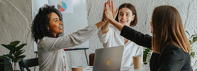 Três mulheres sentadas em uma mesa alegres juntando suas mão ao centro