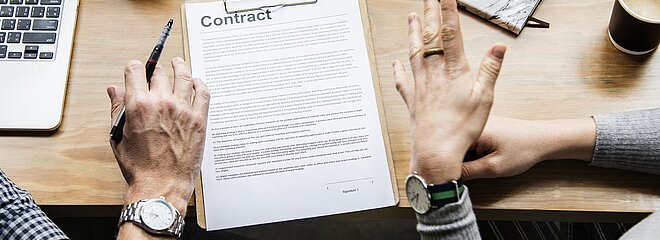 pessoas discutindo em uma mesa com computador e papel onde se lê "contrato" em inglês