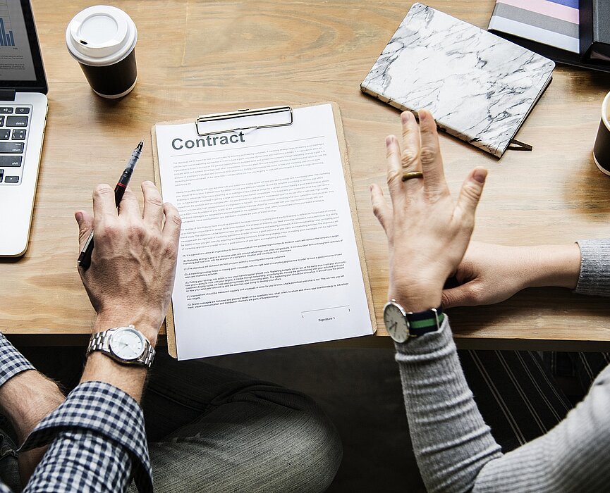 pessoas discutindo em uma mesa com computador e papel onde se lê "contrato" em inglês