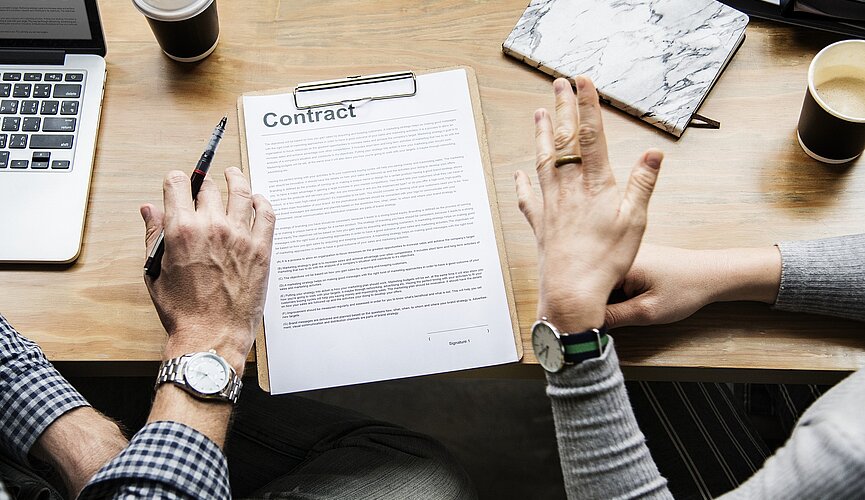 pessoas discutindo em uma mesa com computador e papel onde se lê "contrato" em inglês