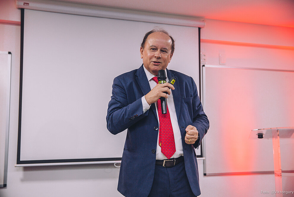 Reitor da UPM discursa em palco com microfone. Painel de projeção na parede ao fundo.
