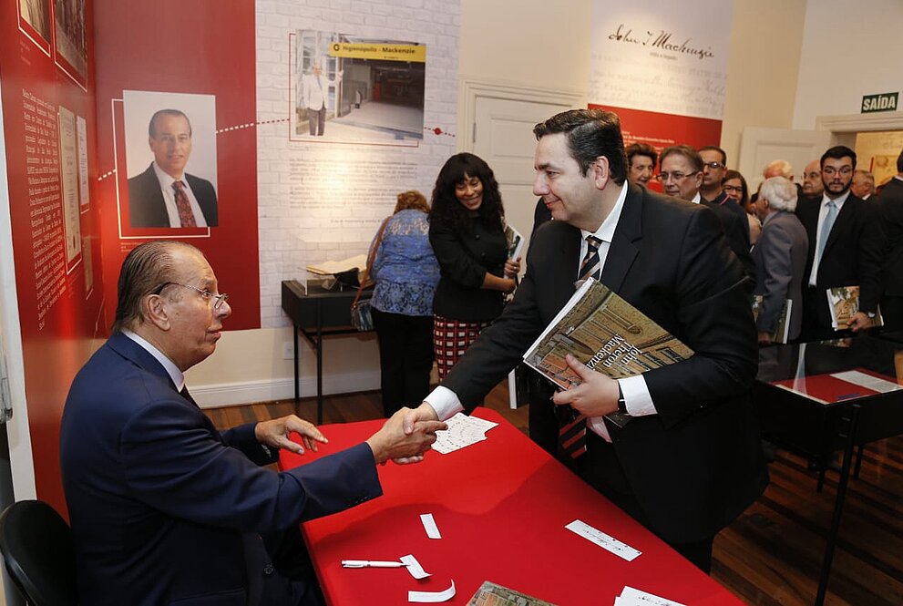 Nelson Câmara sentado à mesa cumprimenta homem que carrega livro no braço.