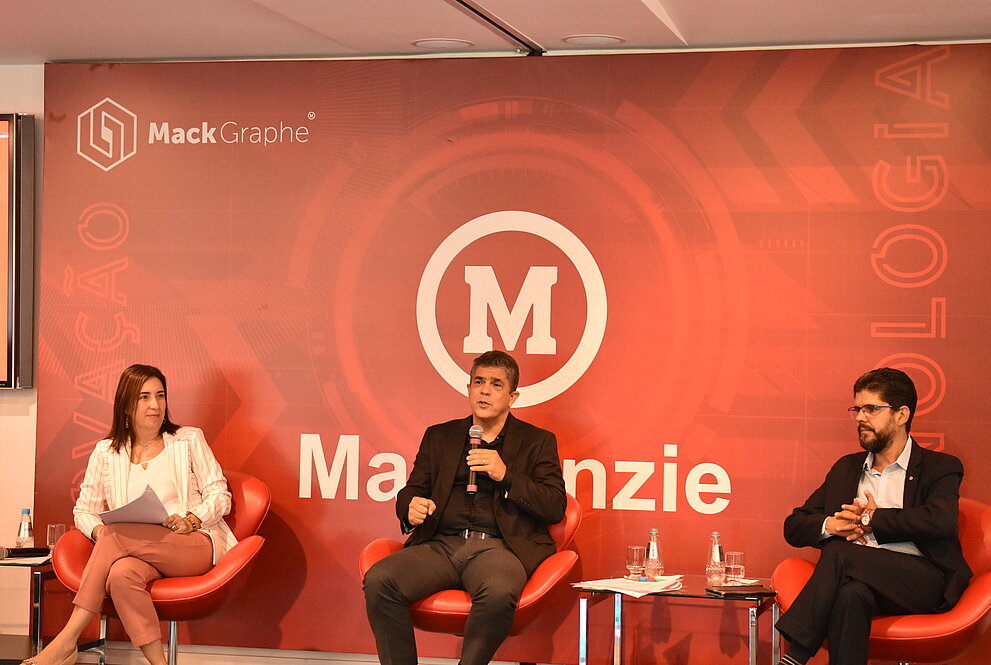 Na foto, da esquerda para direita, estão: Janette, Marco Tullio e Marcelo Coelho. Eles estão respeitando o distanciamento social. Logo atrás, tem um banner do Mackenzie, com a marca e o logo do Mack Graphe.