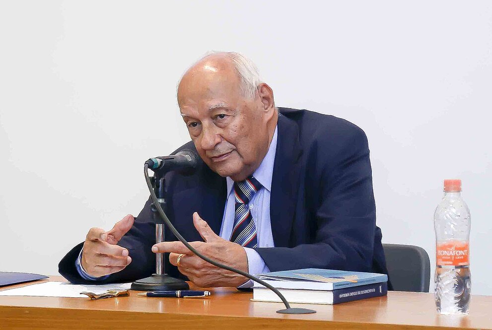 Senhor sentado à mesa, gesticulando com as mãos enquanto fala