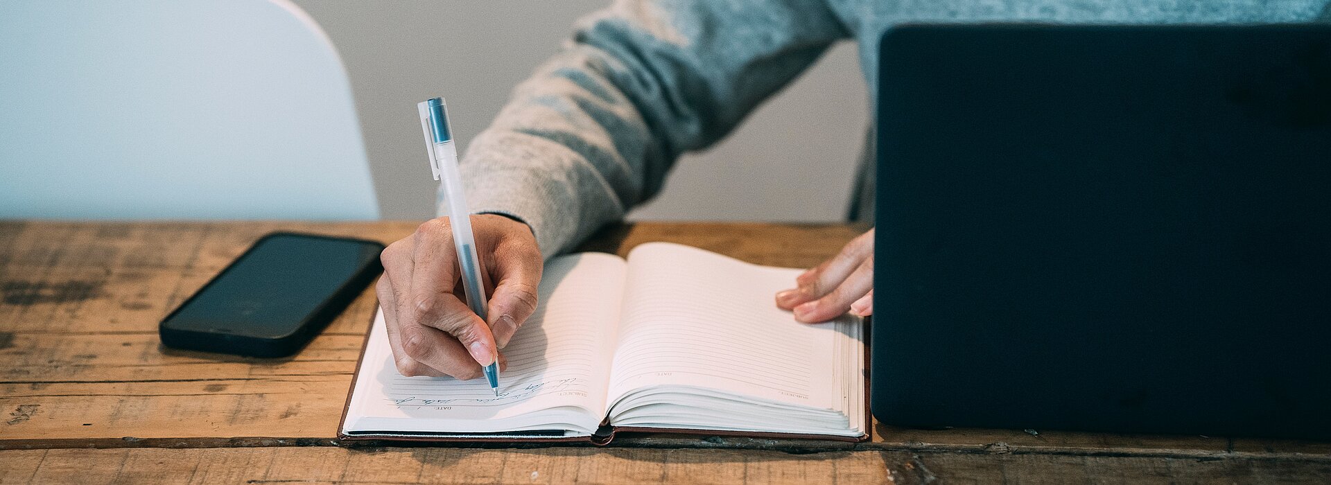 A foto mostra um homem, mas não conseguimos ver seu rosto. Ele está escrevendo em um caderno. Está com um celular do lado e um notebook na sua frente. 