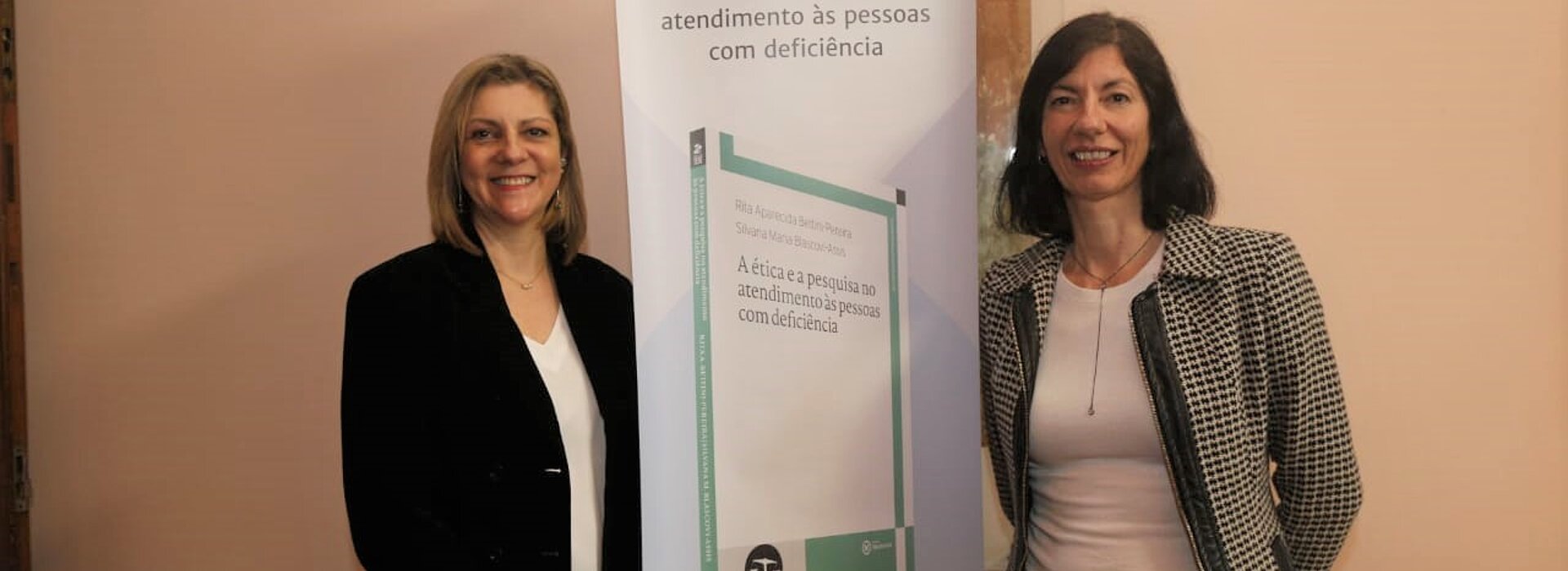 Autoras de pé ao lado do banner de apresentação do livro