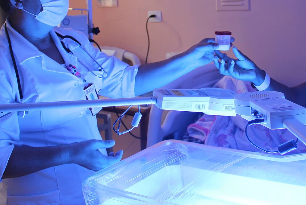 uma mulher usando máscara, luvas e jaleco branco entrega um pote a uma segunda pessoa a qual vemos as mãos com luvas também. O tom da imagem é azulado por conta da iluminação de análise do laboratório