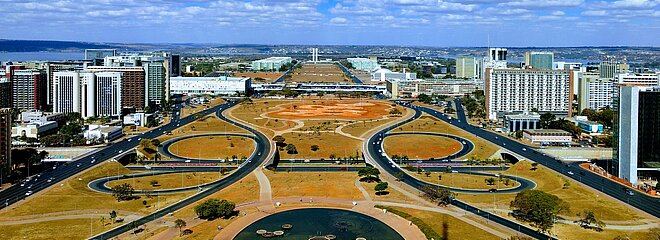 imagem do alto do planalto, em Brasília. o céu está azul, com algumas poucas nuvens brancas e pode-se ver os prédios, ruas e praças