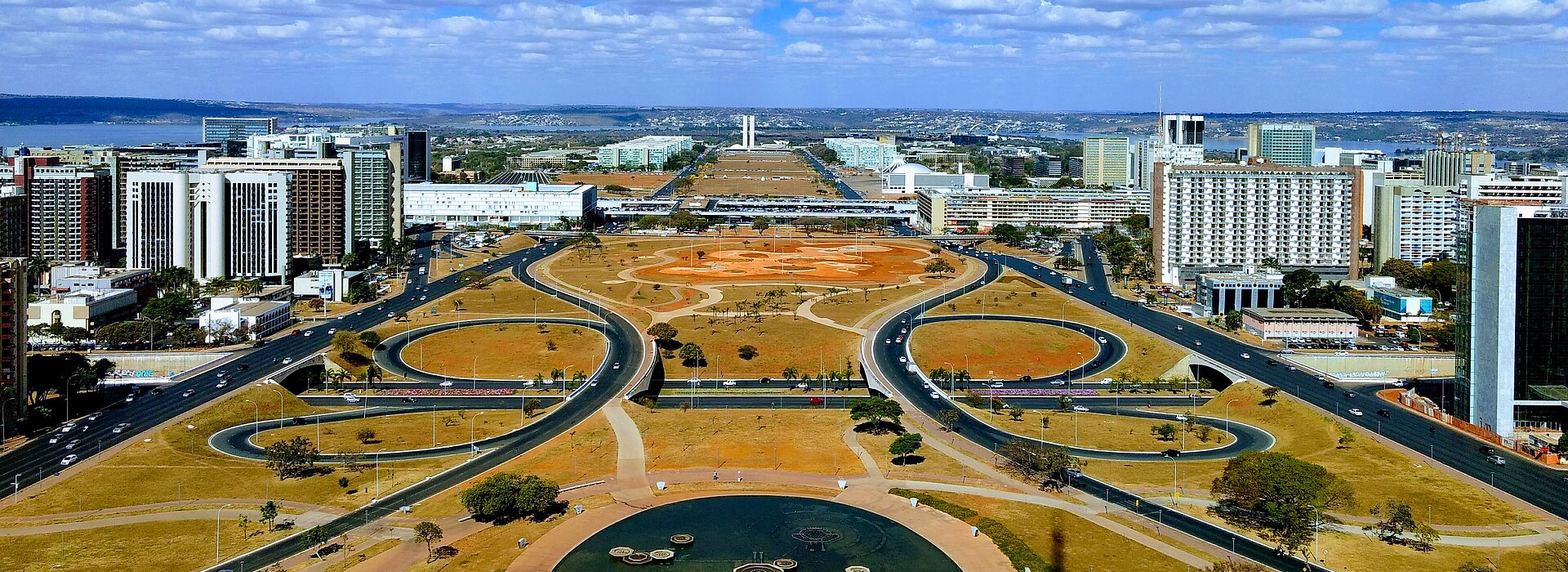 imagem do alto do planalto, em Brasília. o céu está azul, com algumas poucas nuvens brancas e pode-se ver os prédios, ruas e praças