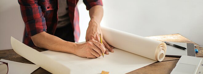 A foto mostra um homem com um lápis na mão começando a desenhar em uma cartolina em branco. Na mesa tem outras folhas e réguas espalhadas. Não conseguimos ver seu rosto. 