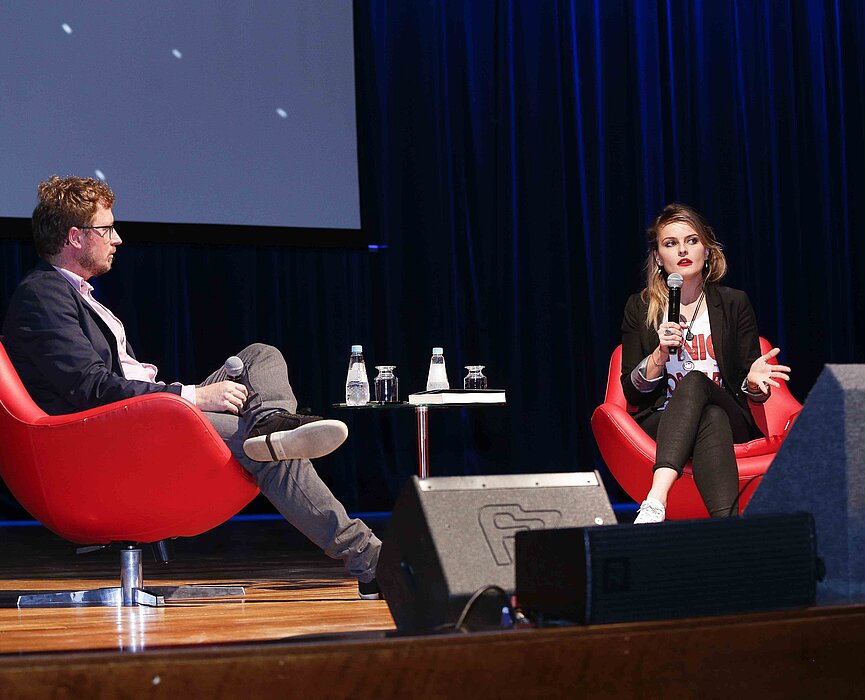 Leandro Narloch e Gloria Álvarez sentados em poltronas vermelhas sobre o palco do auditório Ruy Barbosa.
