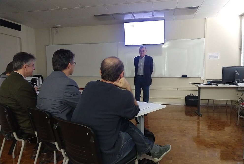 Pessoa idosa em pé fala para outras pessoas sentadas