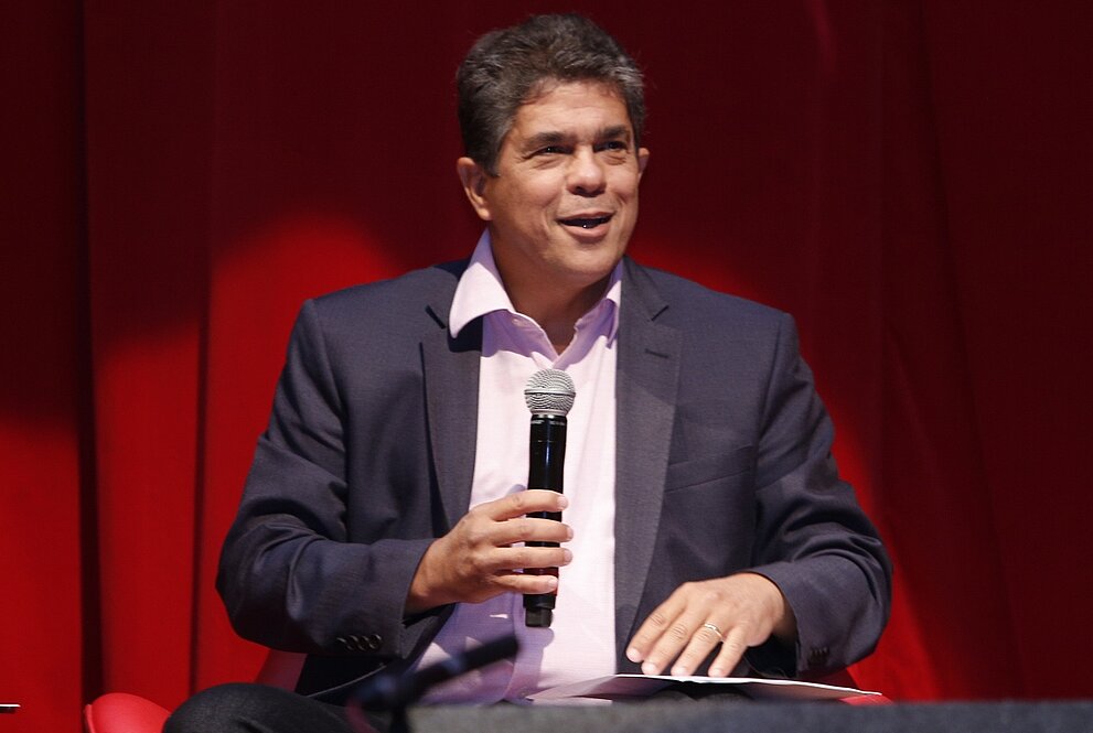 Vicereitor sentado, com microfone em mãos, discursando sobre a Universidade.