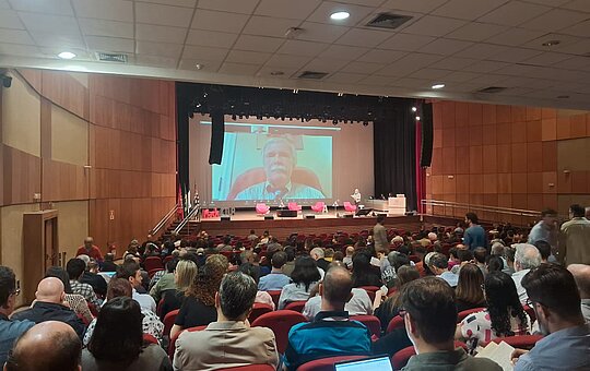 Na imagem, um auditório lotado e uma vídeo sendo passado ao fundo.