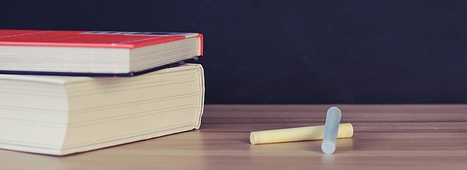 mesa com livros e giz apoiados sobre ela, atrás há um quadro negro