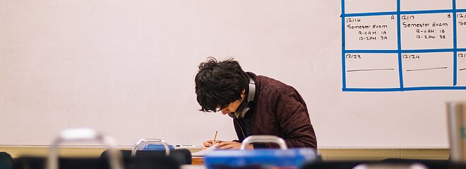 A foto, tirada de longe, mostra menino fazendo uma prova em uma sala de aula. Atrás podemos ver uma lousa. 