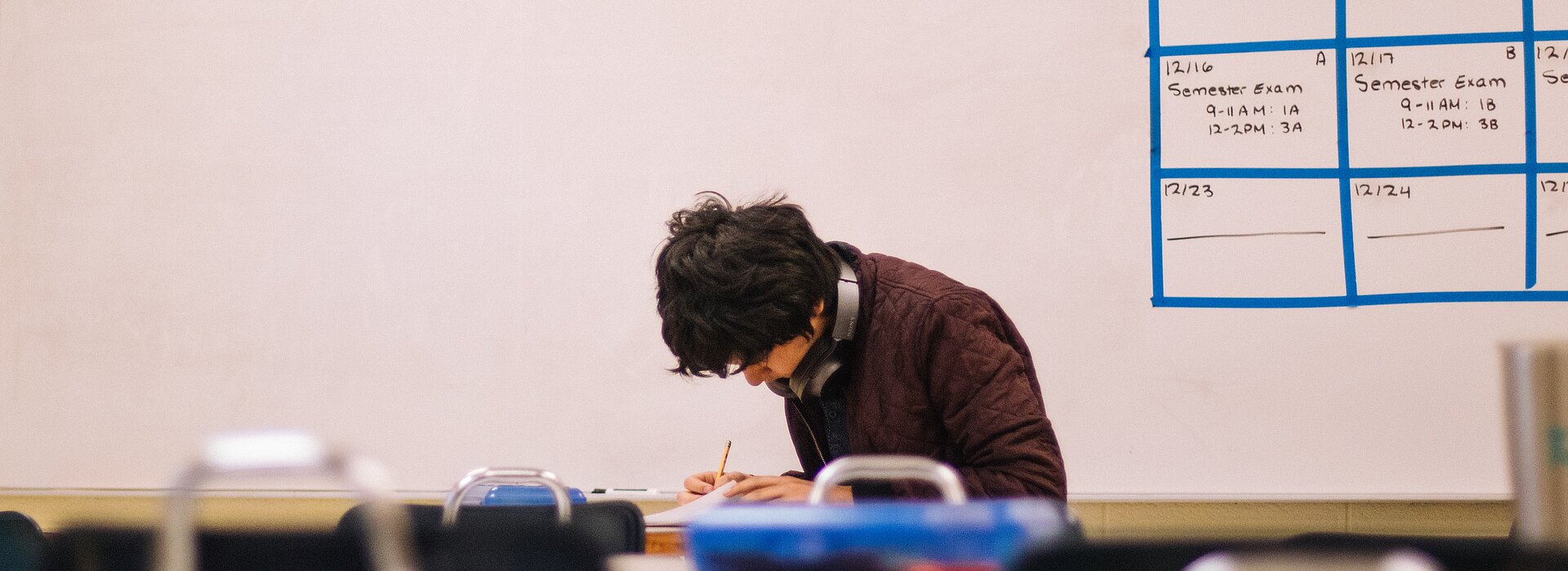 A foto, tirada de longe, mostra menino fazendo uma prova em uma sala de aula. Atrás podemos ver uma lousa. 
