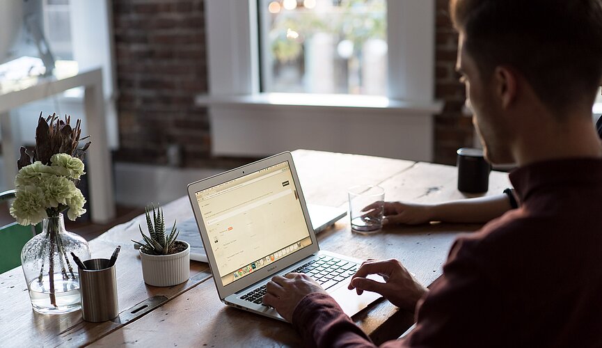 A imagem mostra um homem de lado, mexendo no notebook que está em cima de uma mesa. A imagem não mostra o rosto do homem.