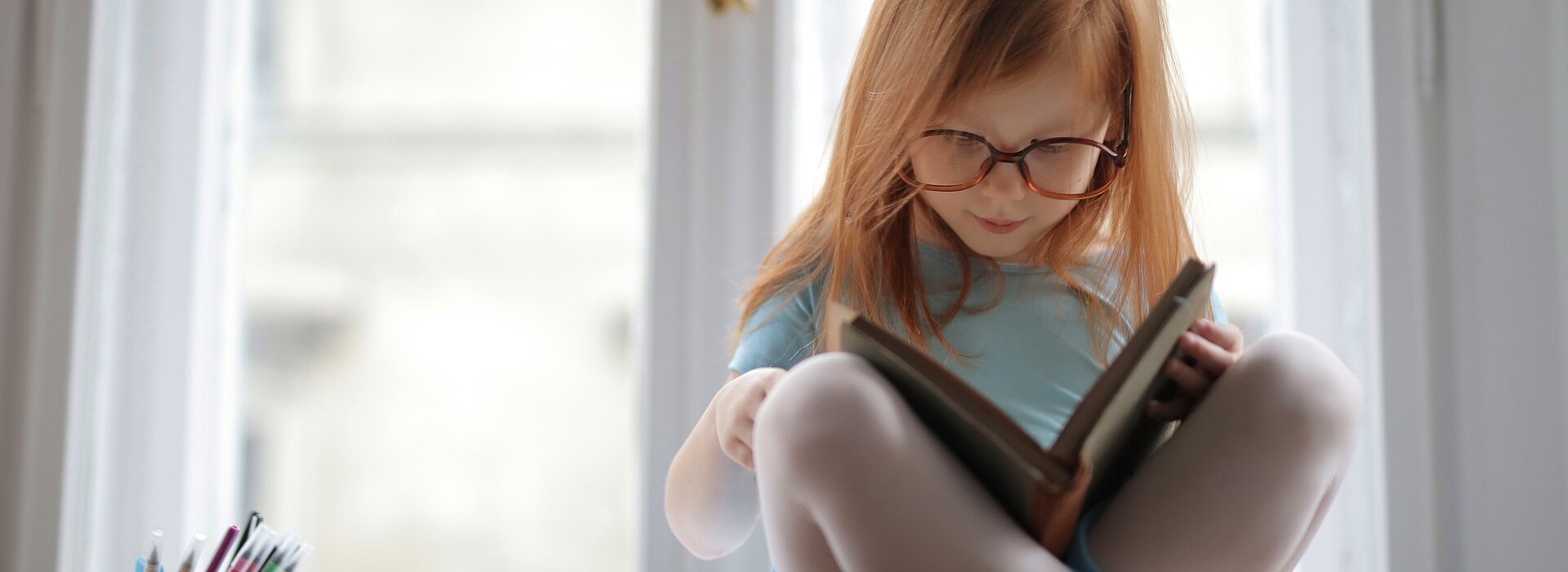 Na foto, uma menina ruiva, de cabelos lisos, está sentada em cima de uma mesa, lendo um livro, com outros livros do lado e muitos lápis em um potinho. A foto foi tirada em frentre a uma janela.