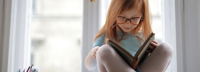 Na foto, uma menina ruiva, de cabelos lisos, está sentada em cima de uma mesa, lendo um livro, com outros livros do lado e muitos lápis em um potinho. A foto foi tirada em frentre a uma janela.