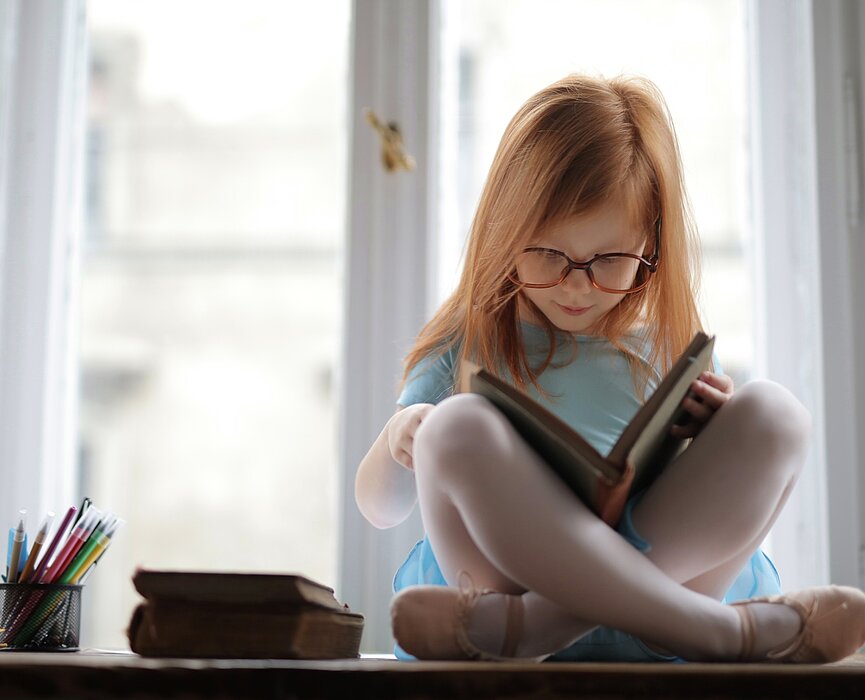 Na foto, uma menina ruiva, de cabelos lisos, está sentada em cima de uma mesa, lendo um livro, com outros livros do lado e muitos lápis em um potinho. A foto foi tirada em frentre a uma janela.