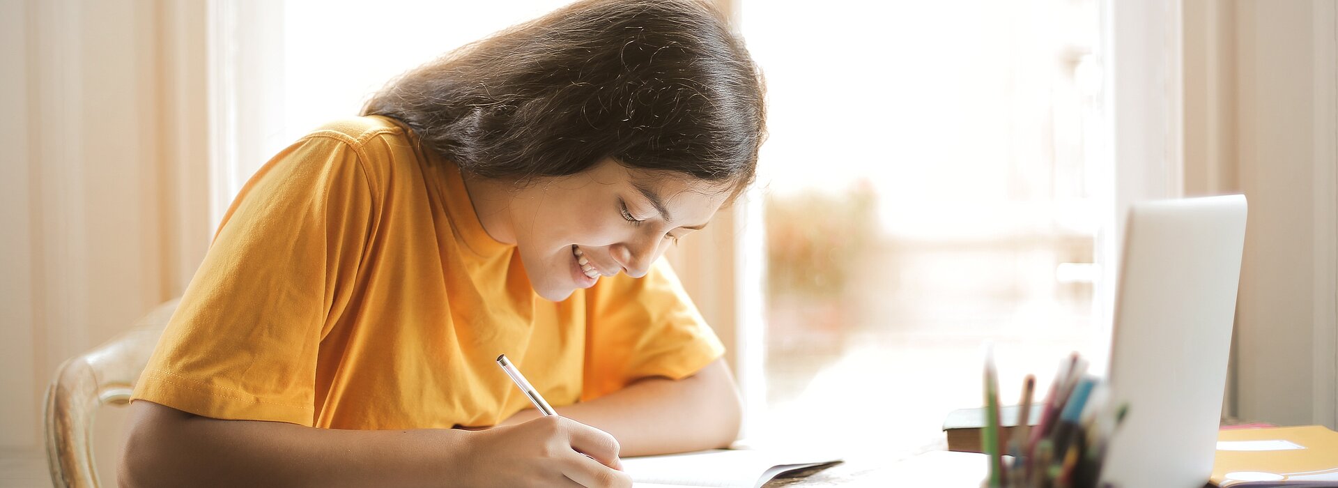Imagem mostra uma menina escrevendo em um folha e um notebook a frente