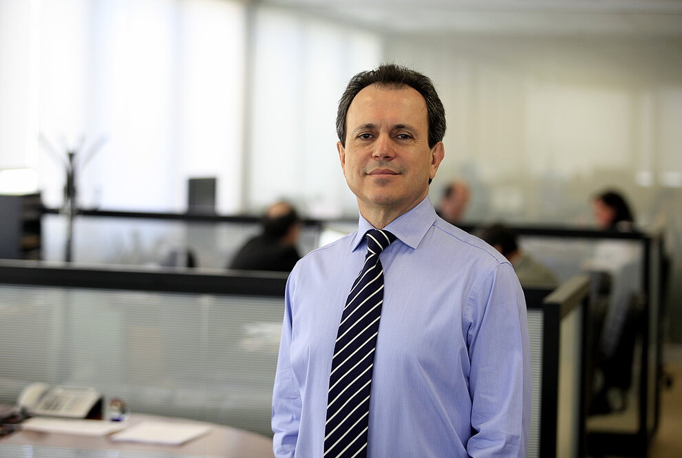 Maurício Meneses de camisa e gravata com escritório ao fundo.