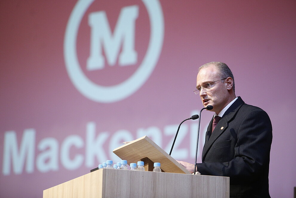 Coordenador do CCSA faz discurso ao púlpito com fundo vermelho com símbolo do Mackenzie
