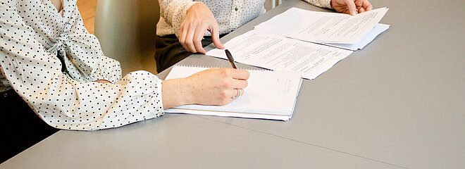 duas mulheres estão em uma mesa, uma delas está assinando um papel e a outra mexendo em outros papéis. as duas parecem estar resolvendo algum negócio. não conseguimos ver seus rostos. 